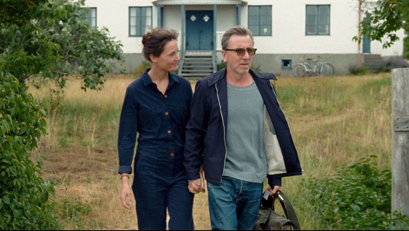 A hand-holding couple walk with a house behind them.