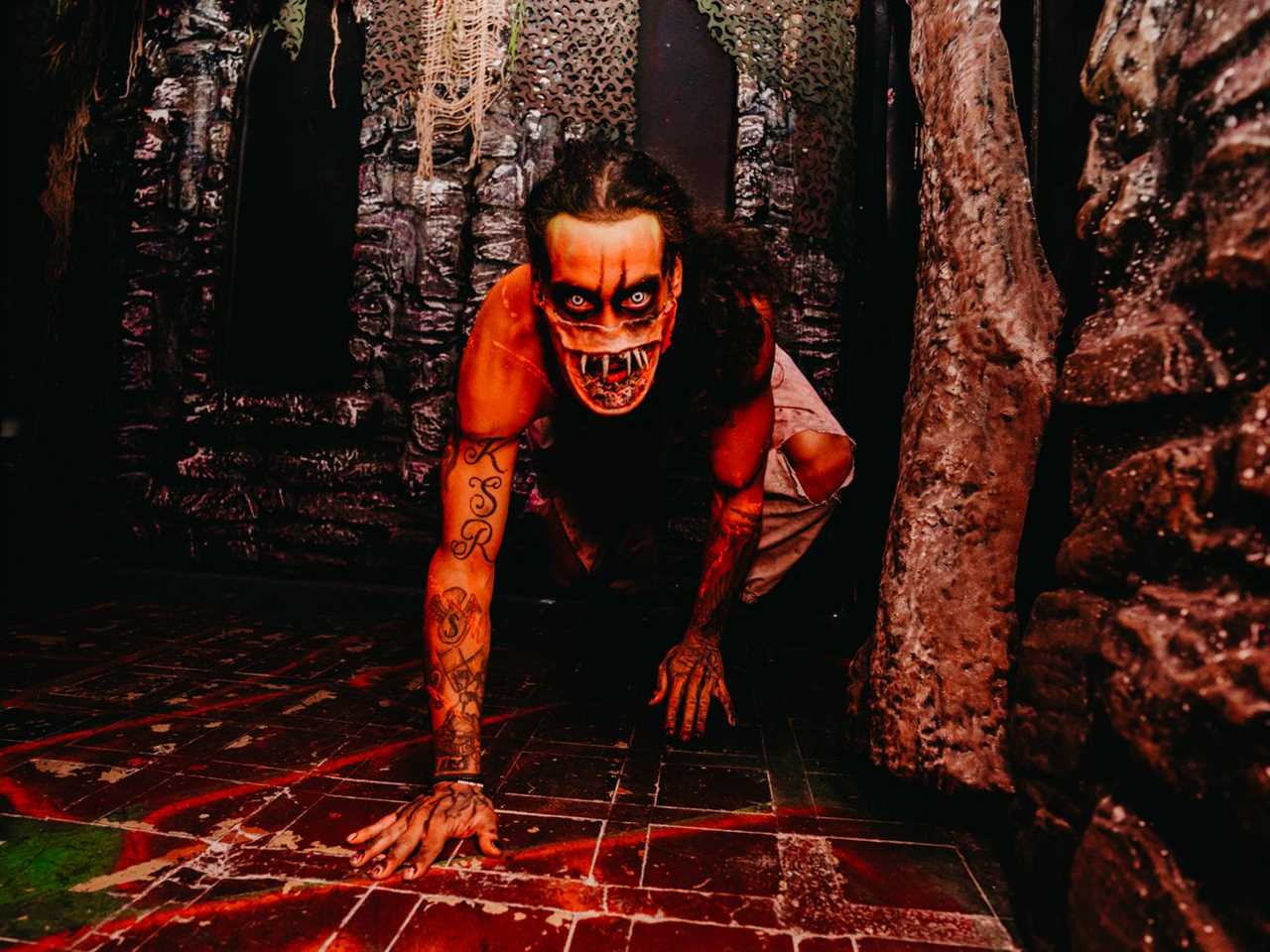 Man in a scary mask crawls towards the camera while bloods spatters on the floor and red lighting illuminates his body. 