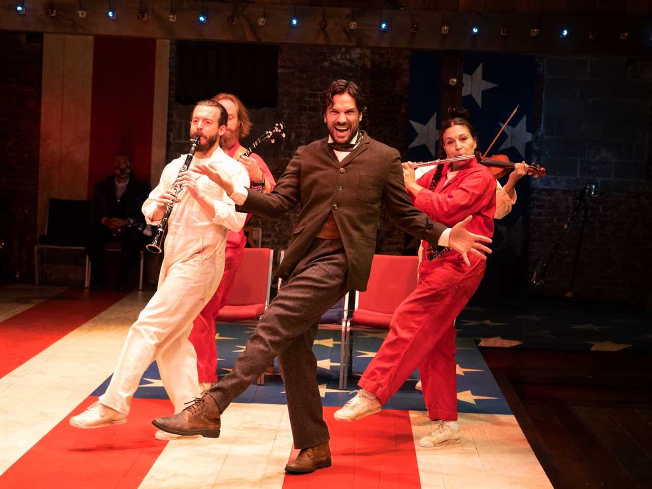 A man in a suit is dancing in front of a trio of musicians. The musicians are wearing prison-style jumpsuits in red, white, and blue. The dancing man in the suit is smiling hugely but his eyes are panicked.