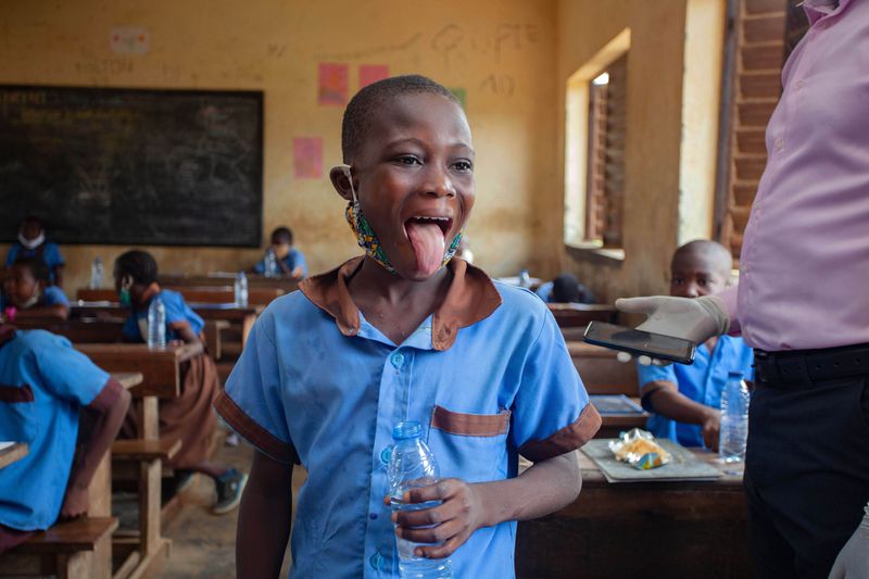 An adult stands next to a child in a classroom full of children at their desks. The standing child is sticking out their tongue.