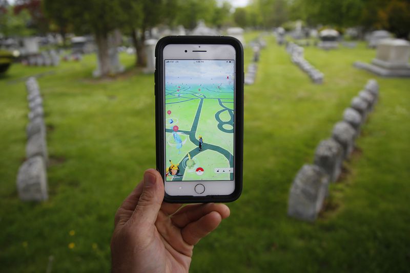 Photo shows a hand holding up an iPhone. On the screen is a still image from the game Pokemon Go, in the background is a cemetery. 