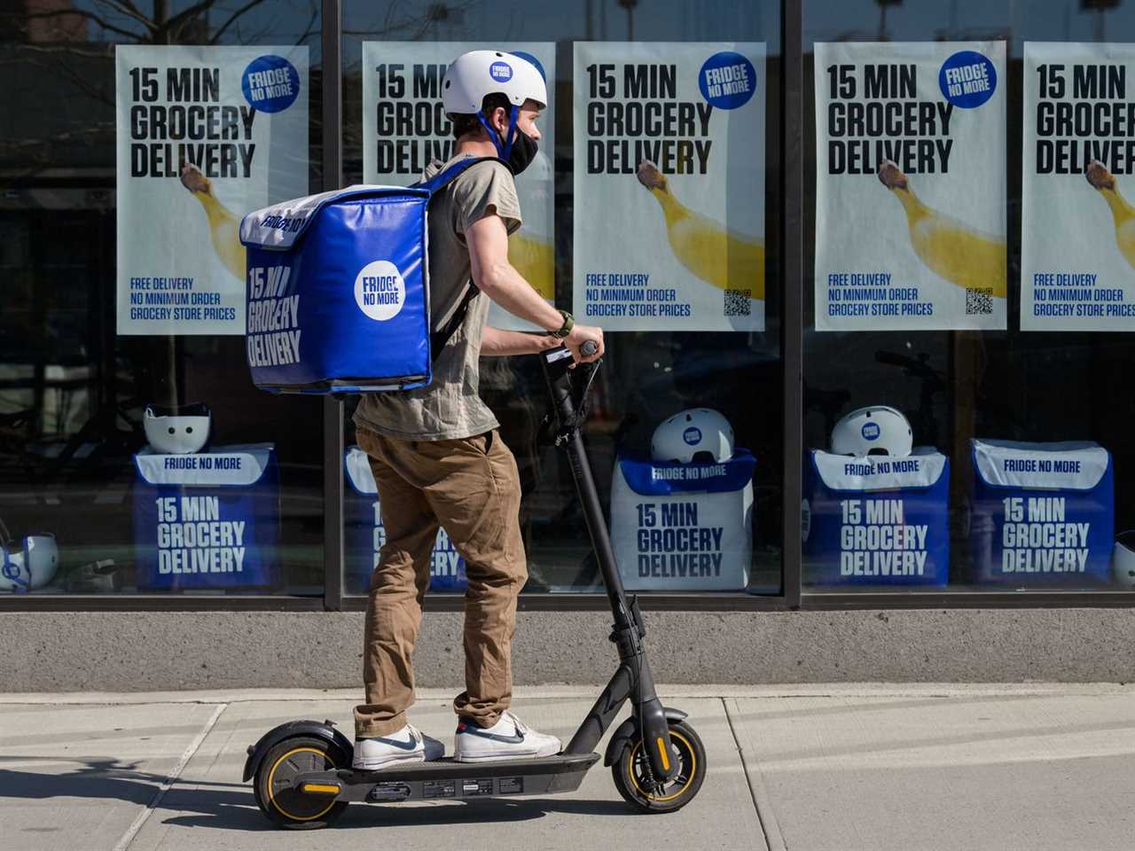 A employee for Fridge No More, an ultrafast delivery service in New York City, rides a scooter on a city sidewalk while carrying a large square pack of groceries on his back.