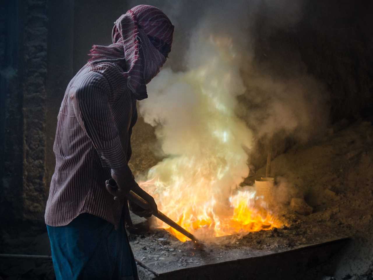 A masked person beside a steaming, glowing vat of molten lead.