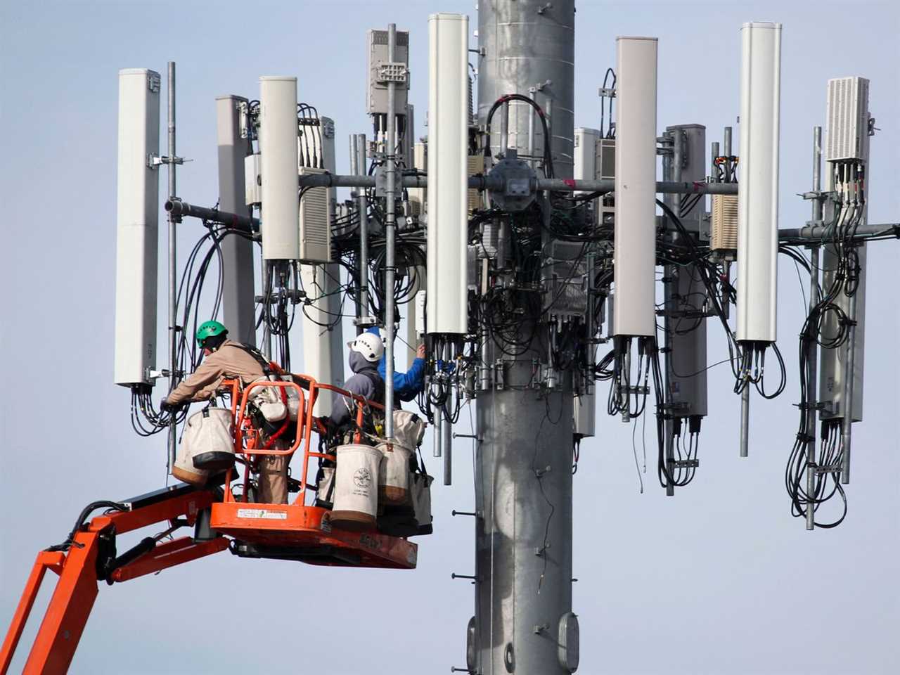 A worker atop a cherry-picker arm reaches out to one of the projecting panels at the top of a huge cellular tower.