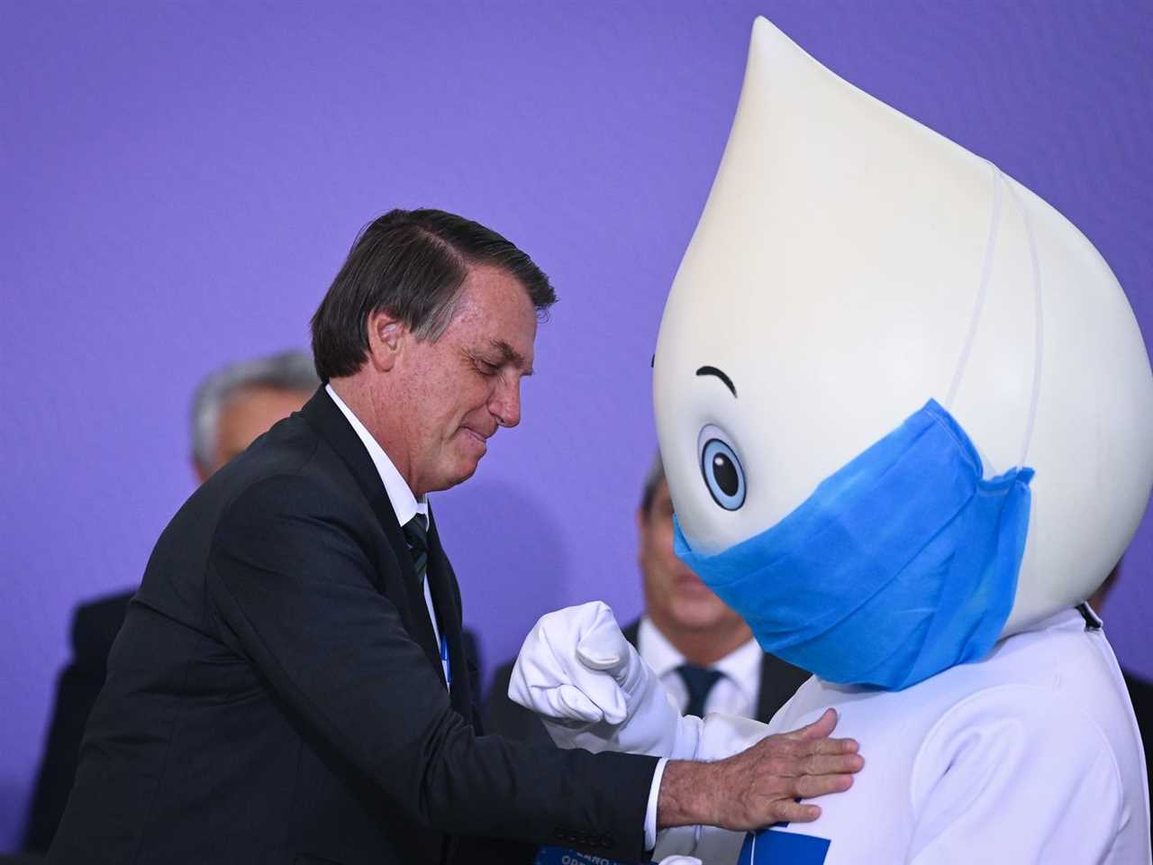 Brazilian president Jair Bolsonaro onstage with a person wearing a raindrop for a head with a huge surgical mask on its face.