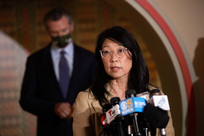 Cynthia Choi speaks over a cluster of media microphones, Gov. Gavin Newsom behind her.