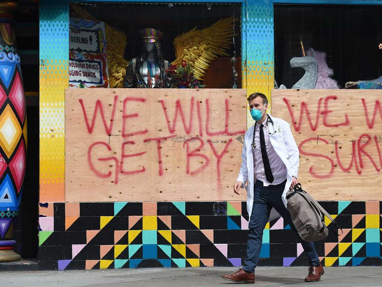 A doctor in a white coat and blue mask walks past a boarded-up shop with spray-painted signs reading “We will get by” and “We will survive.”