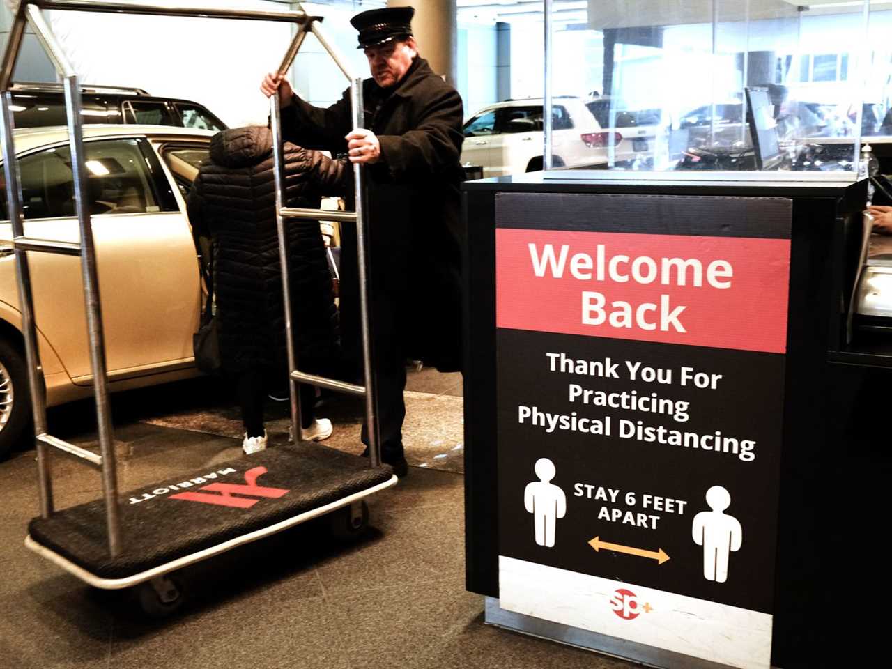 A hotel employee rolls a luggage cart beside a car. A sign beside them reads “Welcome back. Thank you for practicing physical distancing. Stay 6 feet apart.”