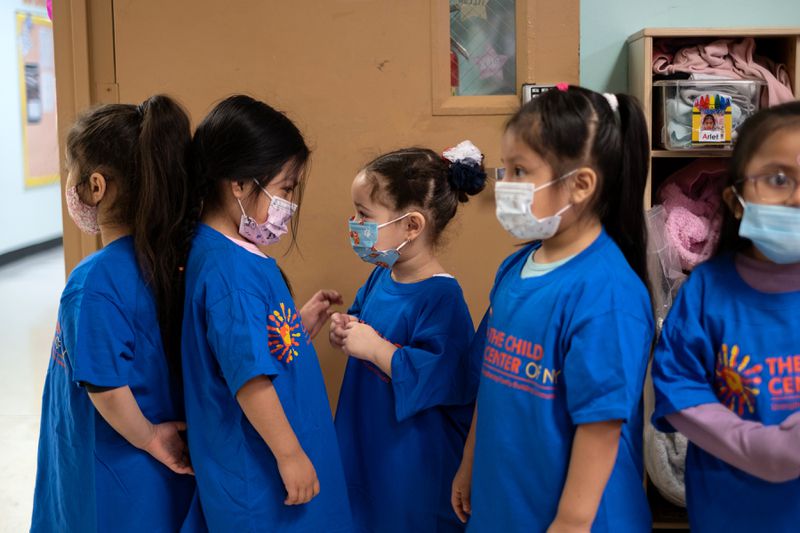 A line of young children in blue T-shirts waits by a sink, talking to each other.