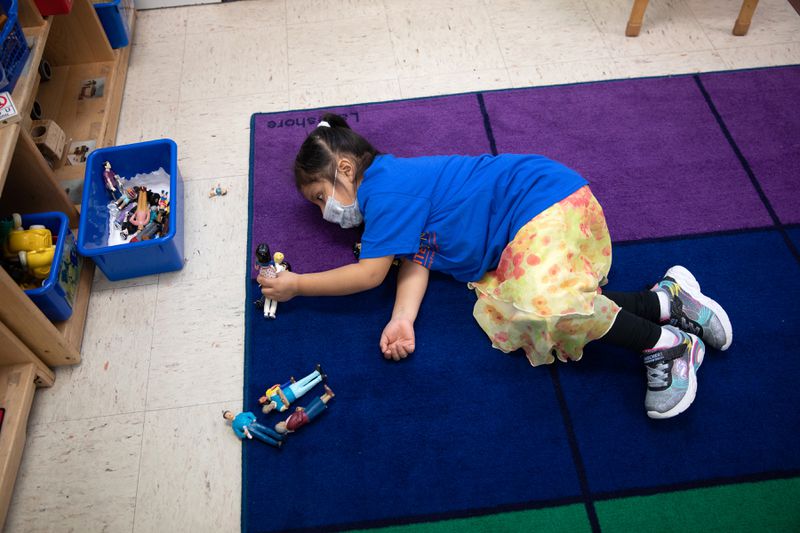 A young girl in a blue T-shirt and yellow skirt lies on a colorful foam mat and plays with a container full of plastic action figures.