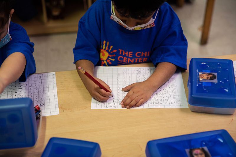 A young girl with a blue T-shirt practices writing letters on a sheet of paper, with a pencil box nearby on the table. 