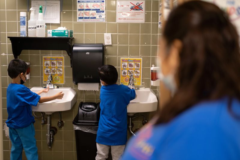 Two young boys in blue T-shirts wash their hands at two separate sinks, watched by their teacher.