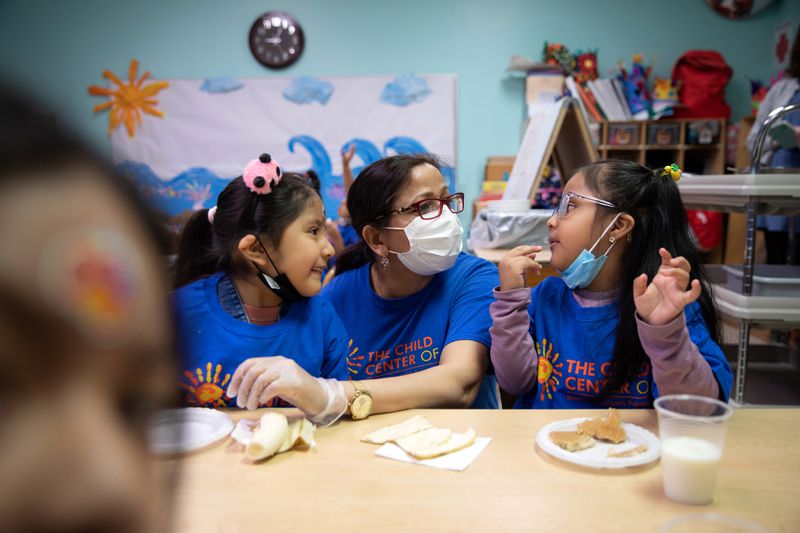 A teacher crouches down at a low table, where two young girls are eating, and talks to them. 