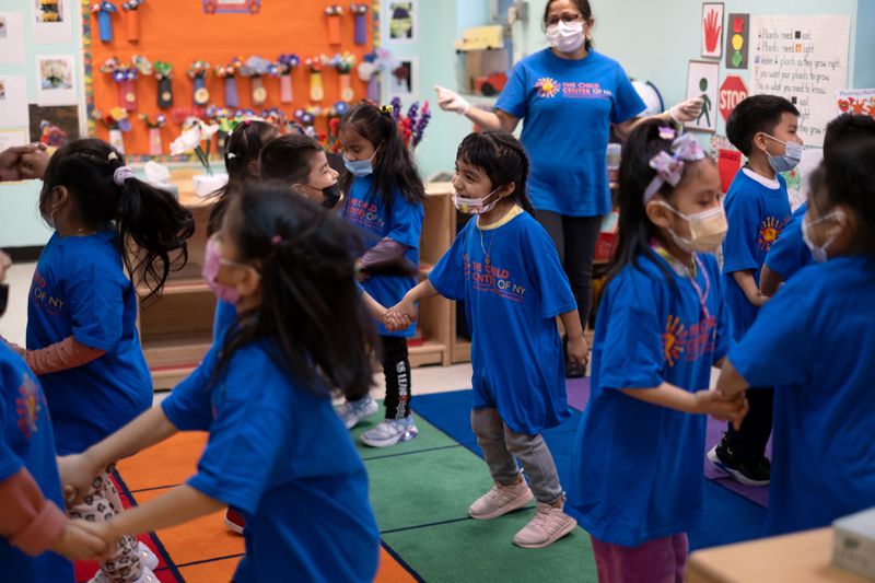 A large group of young children in masks dance and move inside a preschool classroom. 