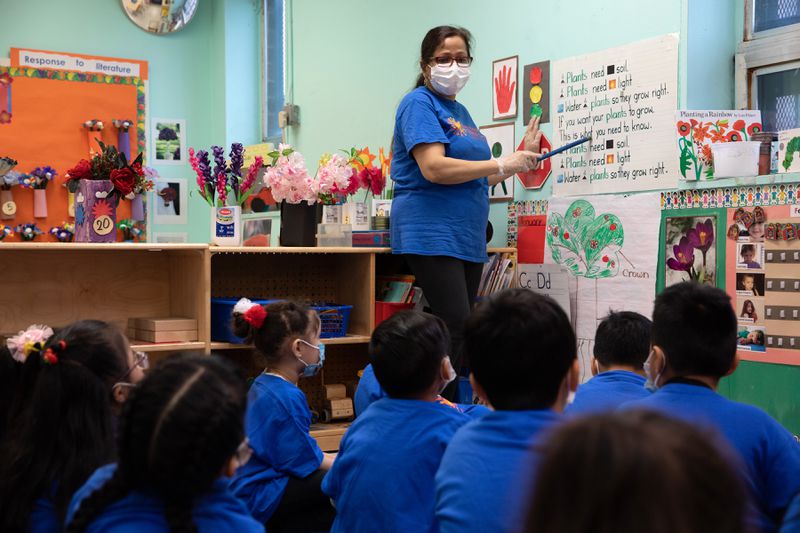 A group of young children, all in blue T-shirts, sits on the floor and listens to their teacher, who stands at the front of the room pointing to words on a large poster on the wall.