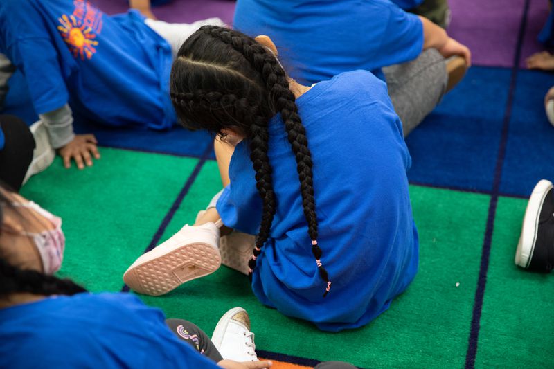 A young girl with two black braids and a blue T-shirt sits with her back to the camera, listening to her teacher and surrounded by her classmates.