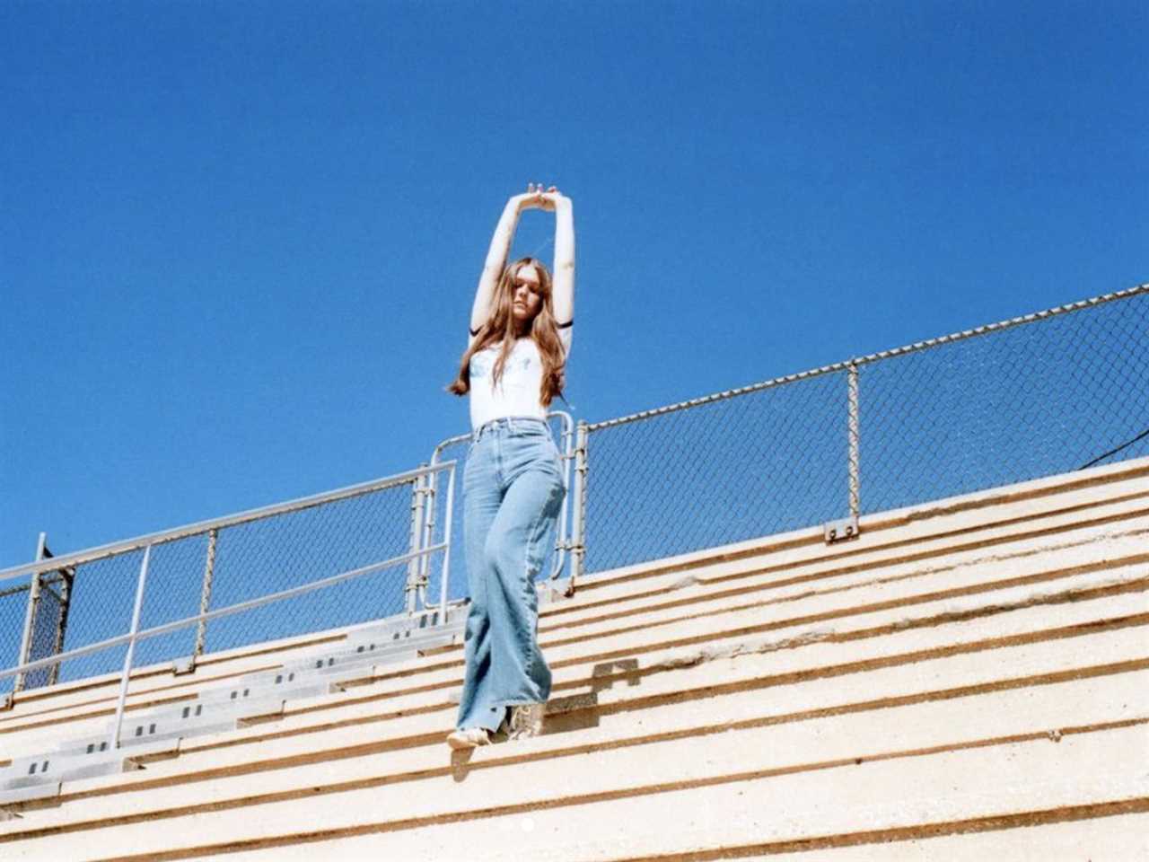 A young woman standing in deserted bleachers with her arms clasped over her head.