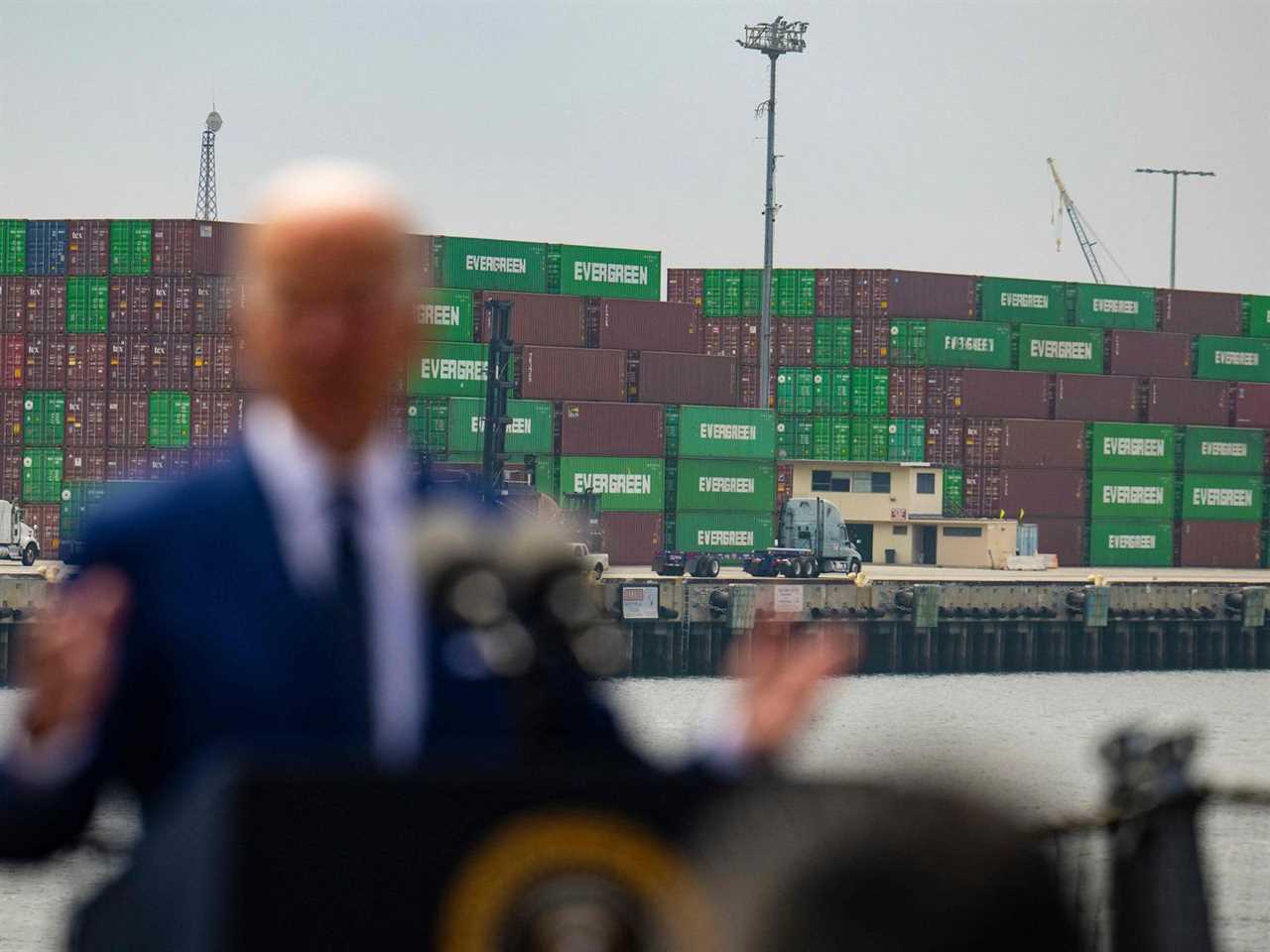 Joe Biden speaks in front of shipping containers, with the containers in focus and Biden blurred.