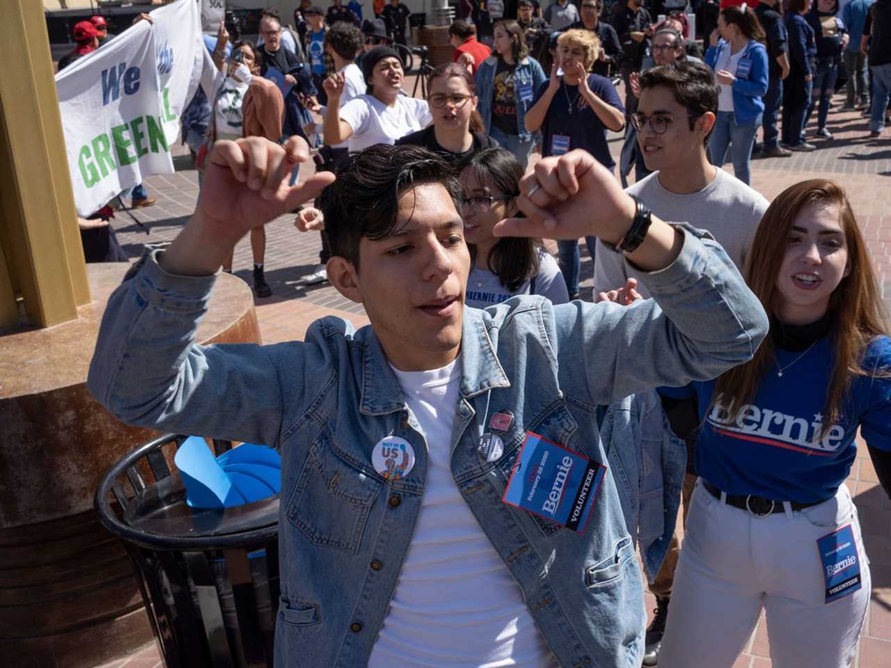 A young Latino man dances with his arms above his head in a blue jean jacket bearing a Bernie Sanders badge; a predominately young, Latino crowd dances in the sunshine behind him.