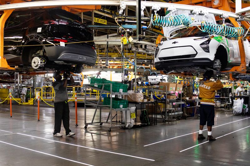 Workers standing on the ground work on cars in an assembly line suspended over their heads.