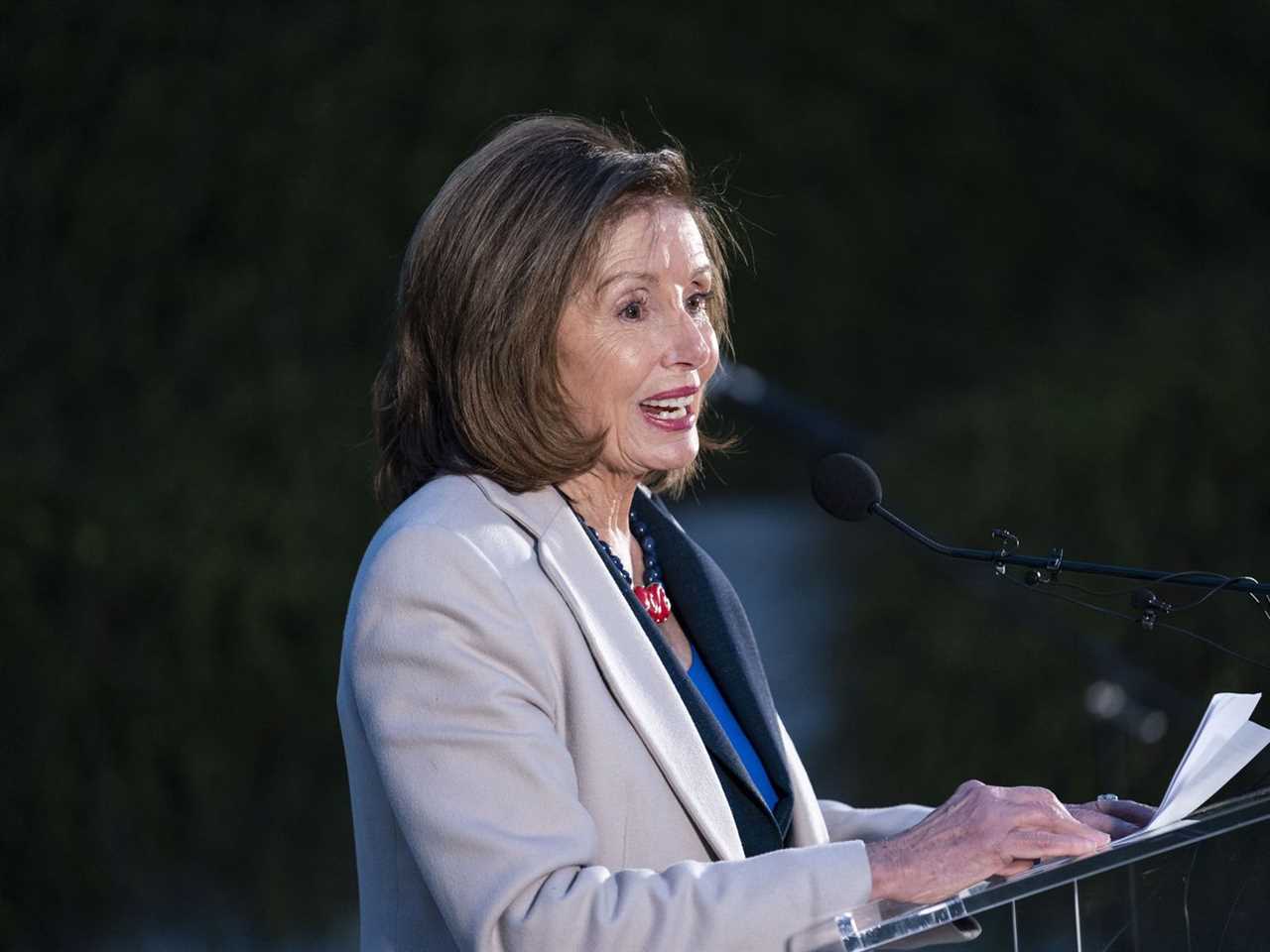 Pelosi, in a pale blue suit, is seen in profile against a dark background, speaking into a microphone. 