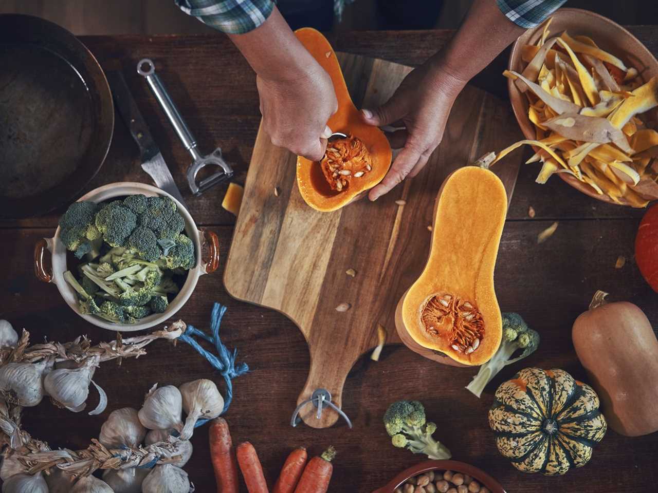 Kitchen table viewed from above strewn with garlic heads, squash, broccoli, and vegetable peelings. Hands visible preparing squash over a cutting board.