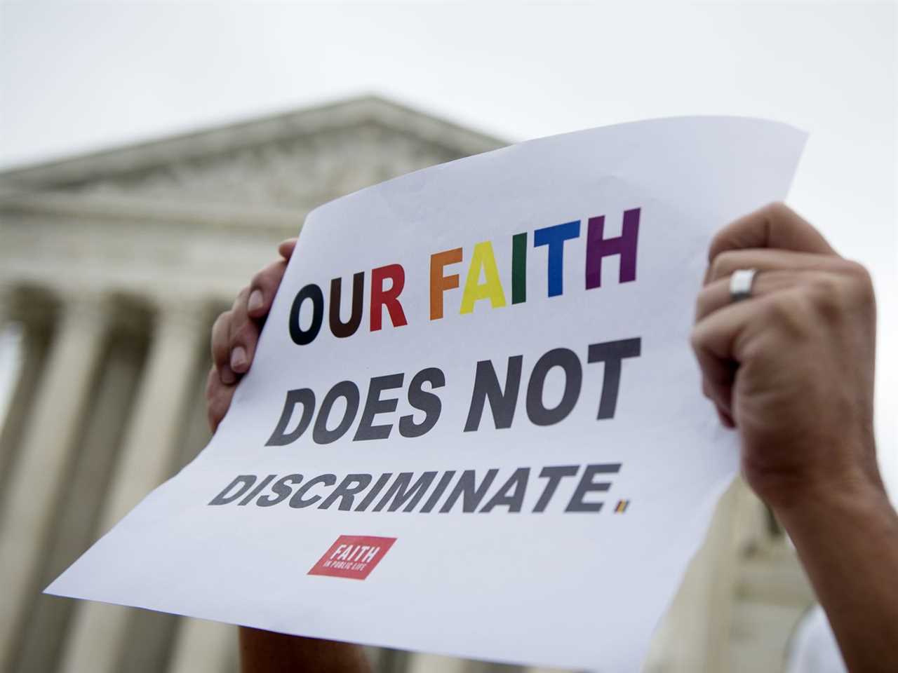 An arm holds up a sign saying “Our faith does not discriminate” in rainbow-colored letters, in front of the US Supreme Court building.