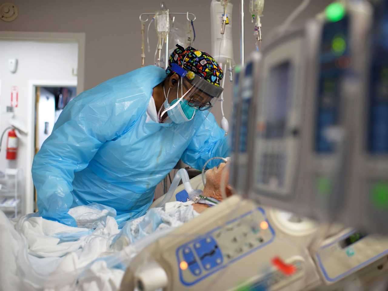 A health care worker in protective mask and gown leans over a patient in a bed in a hospital room. 