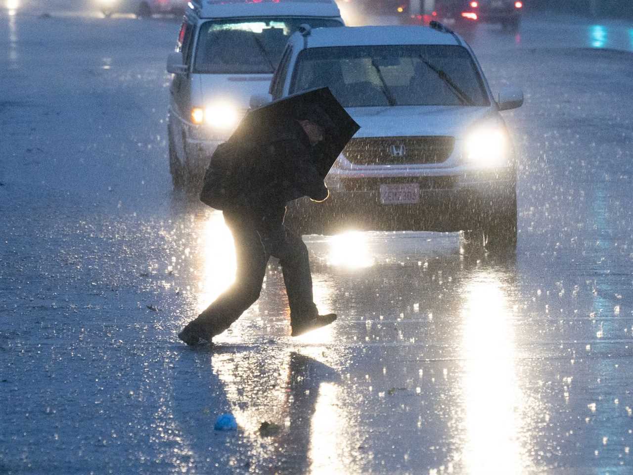 A person carrying an umbrella hunches underneath it as they cross the street under what looks like heavy rain. Cars with their windshield wipers and headlights turned on wait in the background.