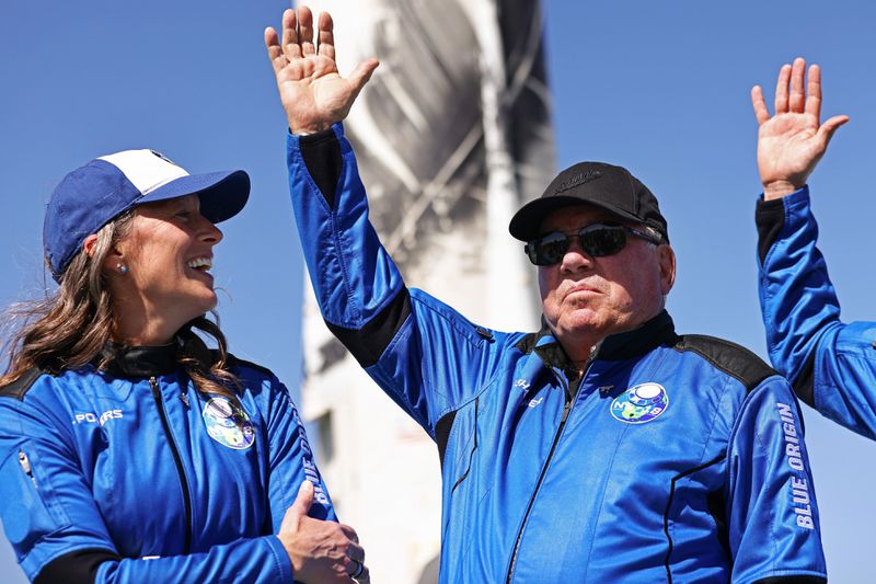 William Shatner, dressed in a blue flight jacket, sunglasses, and baseball cap, waves. 