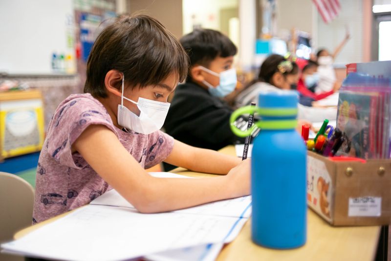 Children in masks work on a long table in a brightly lit kindergarten classroom.