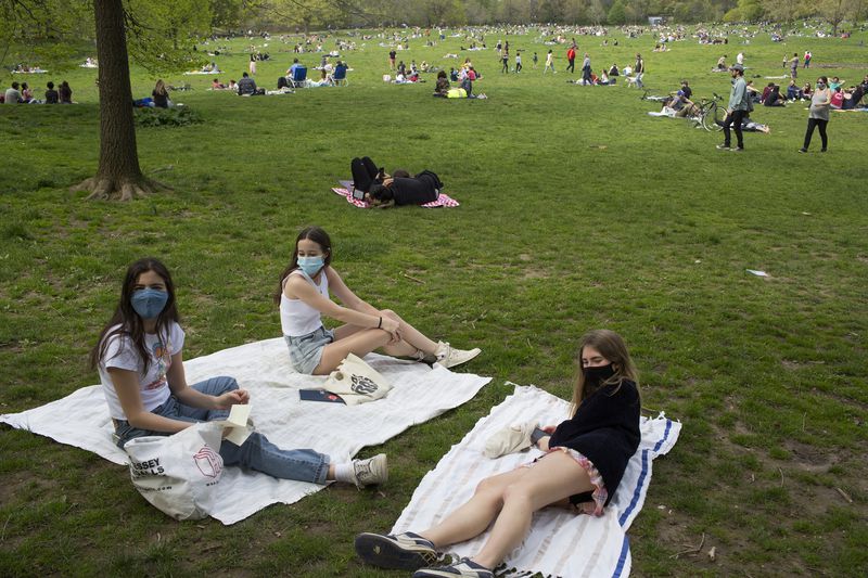 Three teens sit on blankets on grass in a park. They’re wearing masks.