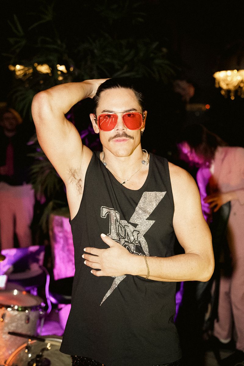 Tom Sandoval wears orange sunglasses, a black tank top with a silver lightning bolt, and metal jewelry, while posing with one arm above his head at a party. 