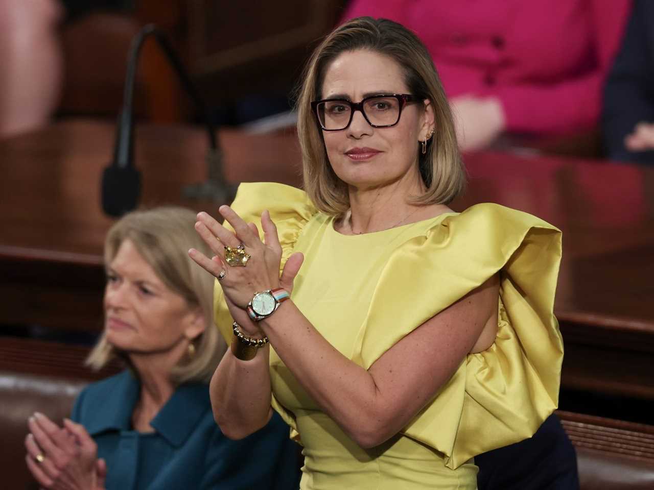 Sen. Sinema, wearing a shiny yellow dress with puffy sleeves, applauds.