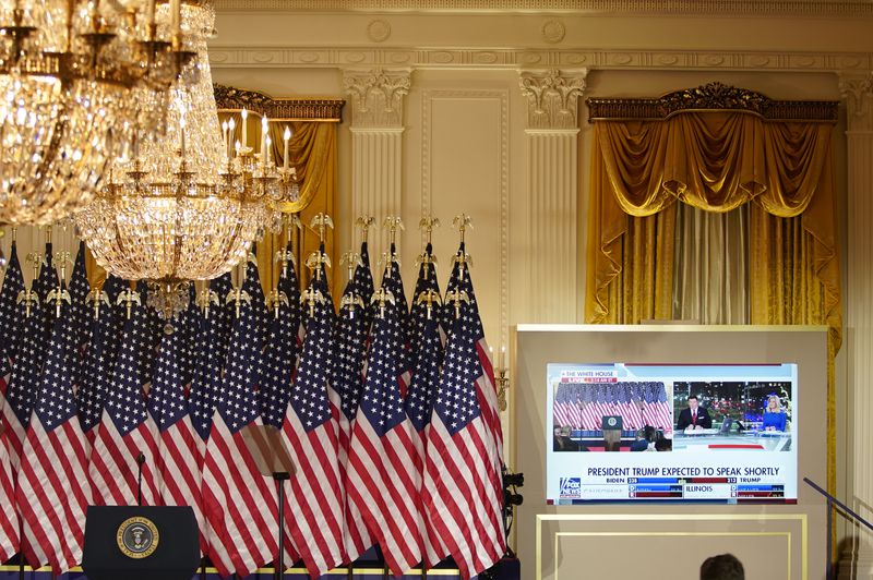 To the right of a row of US flags, a podium bearing the seal of the president, and beneath massive chandeliers, a flatscreen television plays Fox News’ 2020 election programming. 