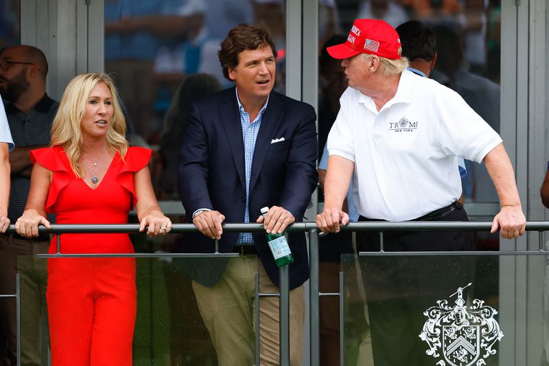 Greene, in a bright red jumpsuit looks out over a balcony, as Carlson in a navy sports jacket, blue shirt, and tan slacks, holds a bottle of Perrier. Trump, in a white polo bearing his golf club’s logo and a red MAGA hat, is seen in profile, speaking to the others.