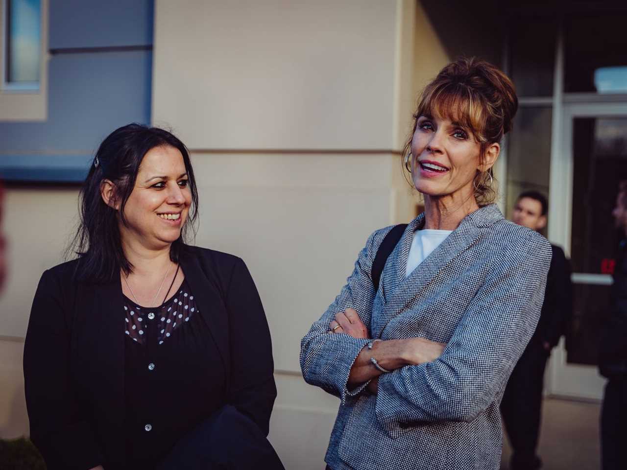 Two women dressed in business attire smile and appear relaxed, outside a concrete-walled building.