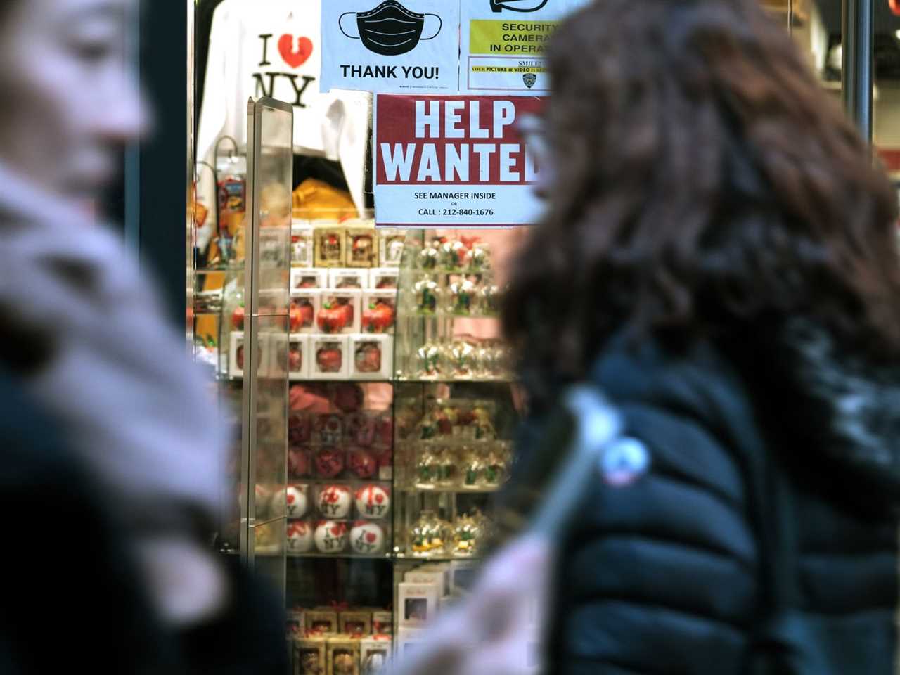 Pedestrians on the sidewalk in front of a glass shop front with a help wanted sign in the window.