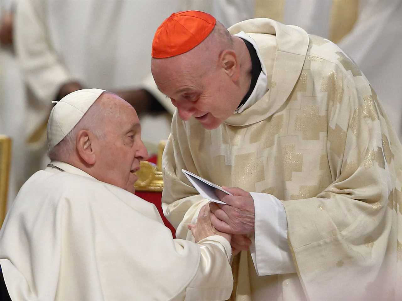 The Catholic pope, seated and wearing a white robe and cap, speaks with a bishop, standing and leaning in, wearing a white robe and red cap.