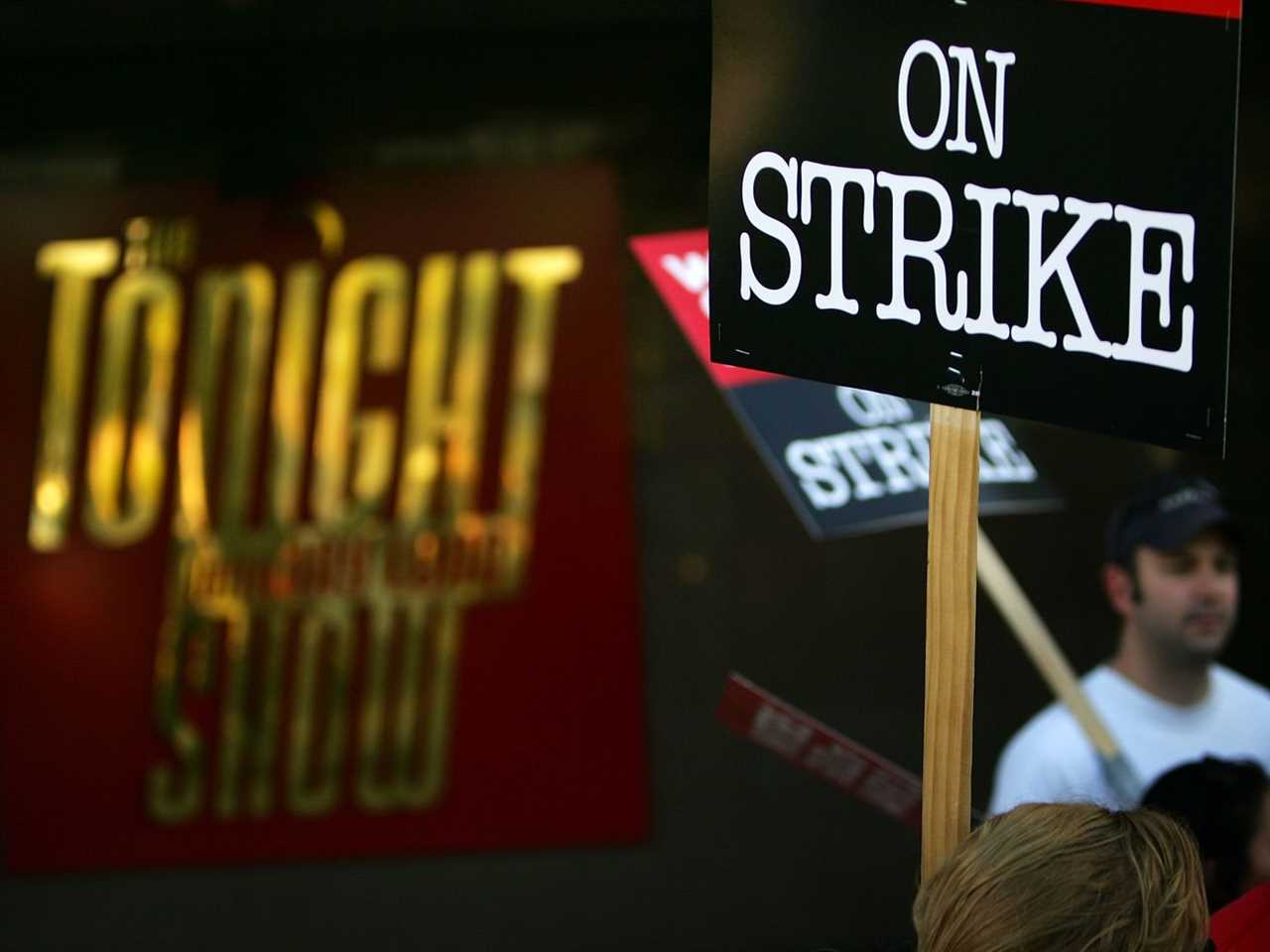 A man in a white shirt holds a sign reading “On strike” in front of a theater with the Tonight Show logo on it.