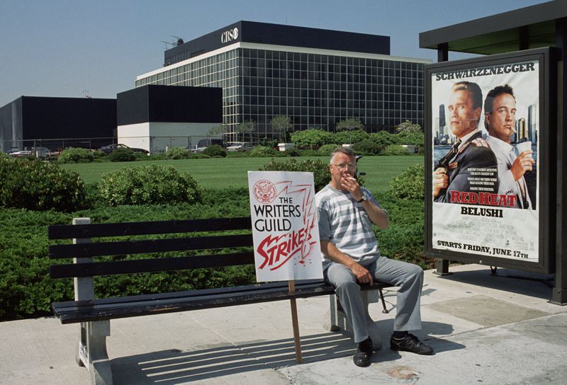 A man in light summer clothes sits at a bus stop in Los Angeles, holding a Writers Guild strike sign and smoking a cigarette.