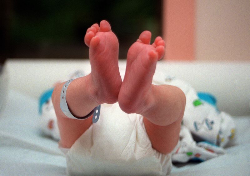 A newborn with a hospital tag on its leg, lying down, seen from below with bare feet and a diapered butt.