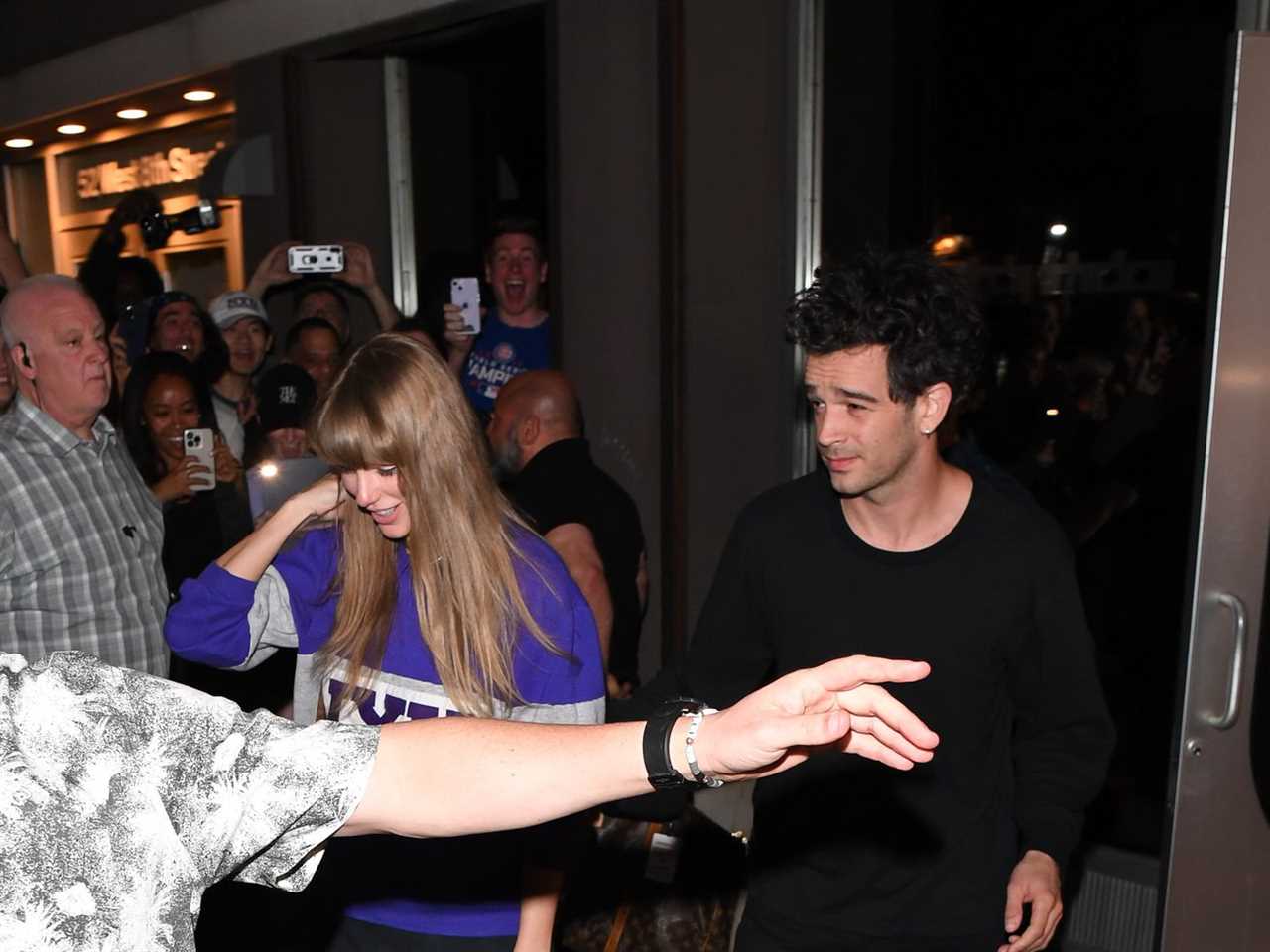 A photo shows Taylor Swift and Matty Healy exiting a glass door onto a crowded sidewalk, side by side. A person otherwise unseen is throwing an arm in front of the pair to keep onlookers away.