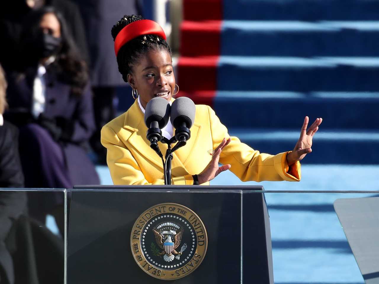 Gorman, a young Black woman in a red hat and yellow suit, gestures as she recites a poem from a podium with the presidential seal.