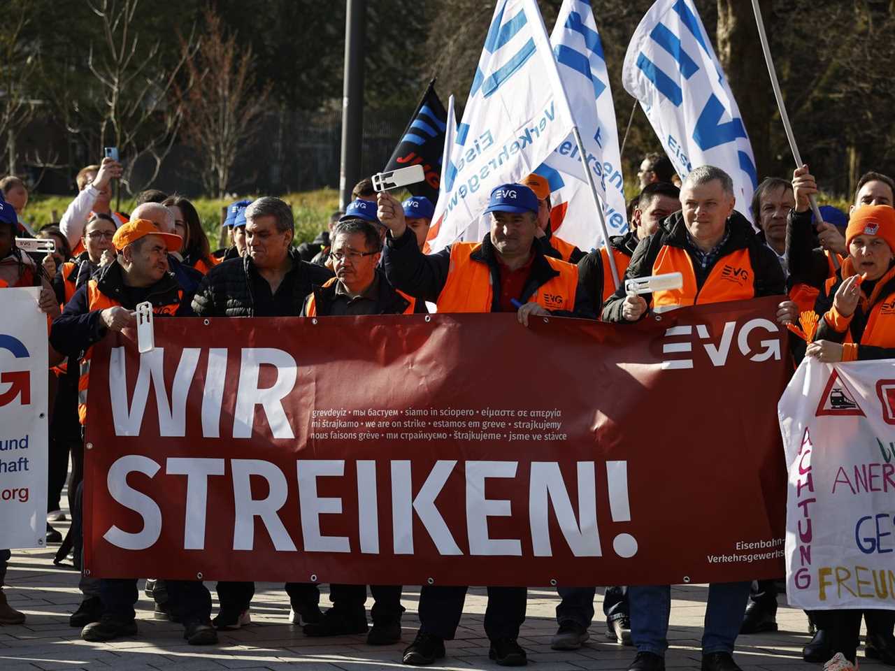 A crowd of protesters wearing orange vests hold signs. 