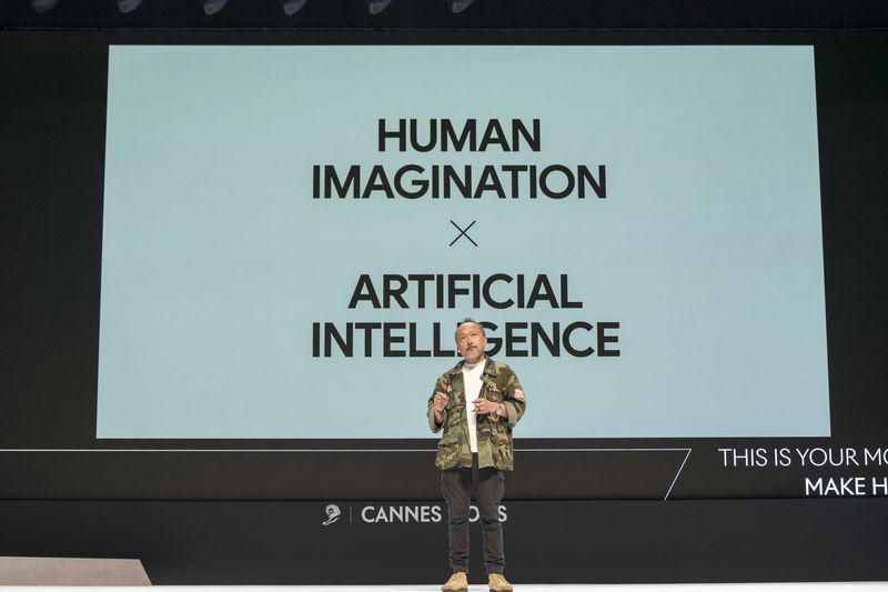 A presenter onstage in front of a screen that reads “Human intelligence x artificial intelligence.”