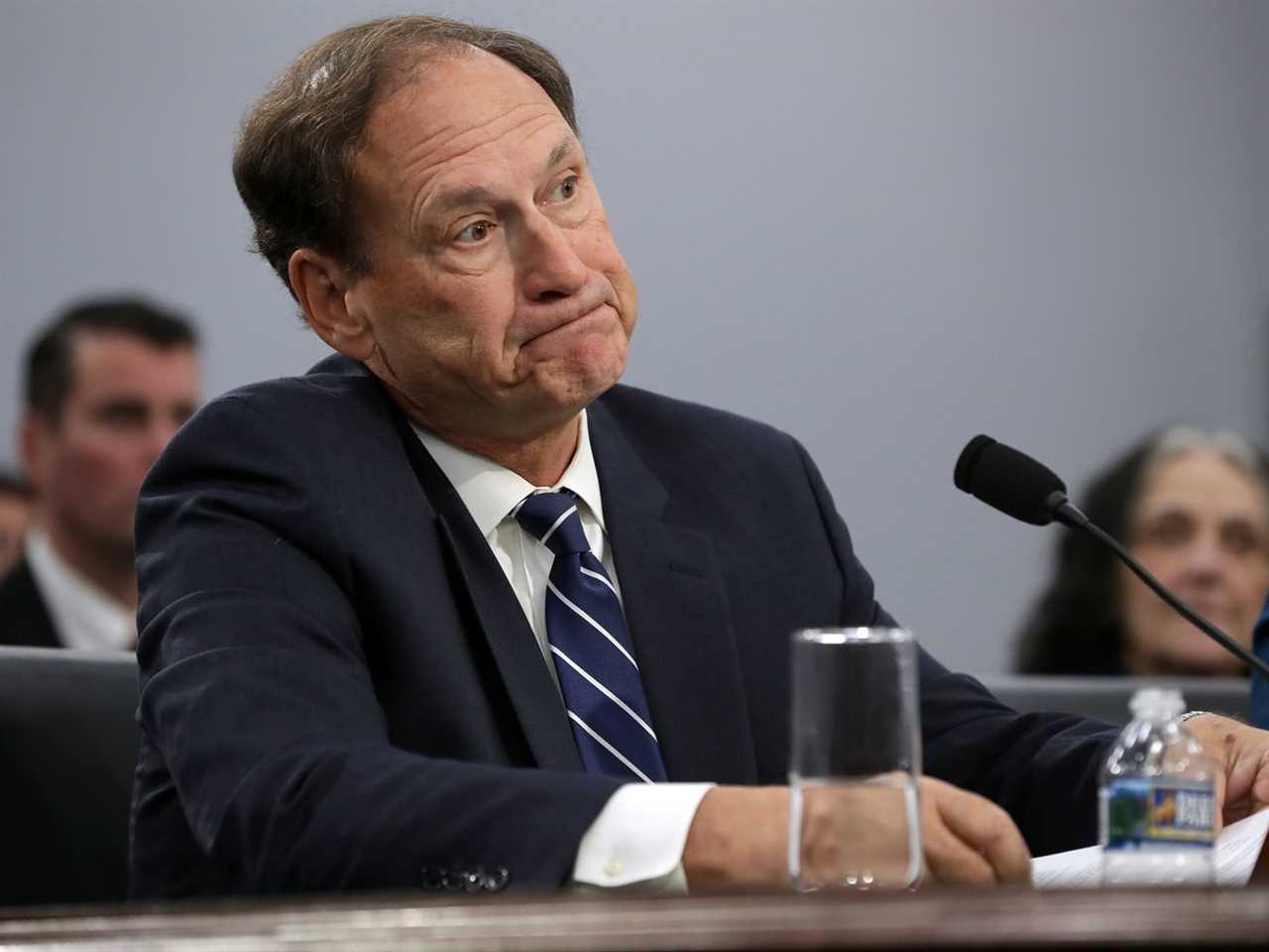 Samuel Alito sits at a desk facing a microphone. 