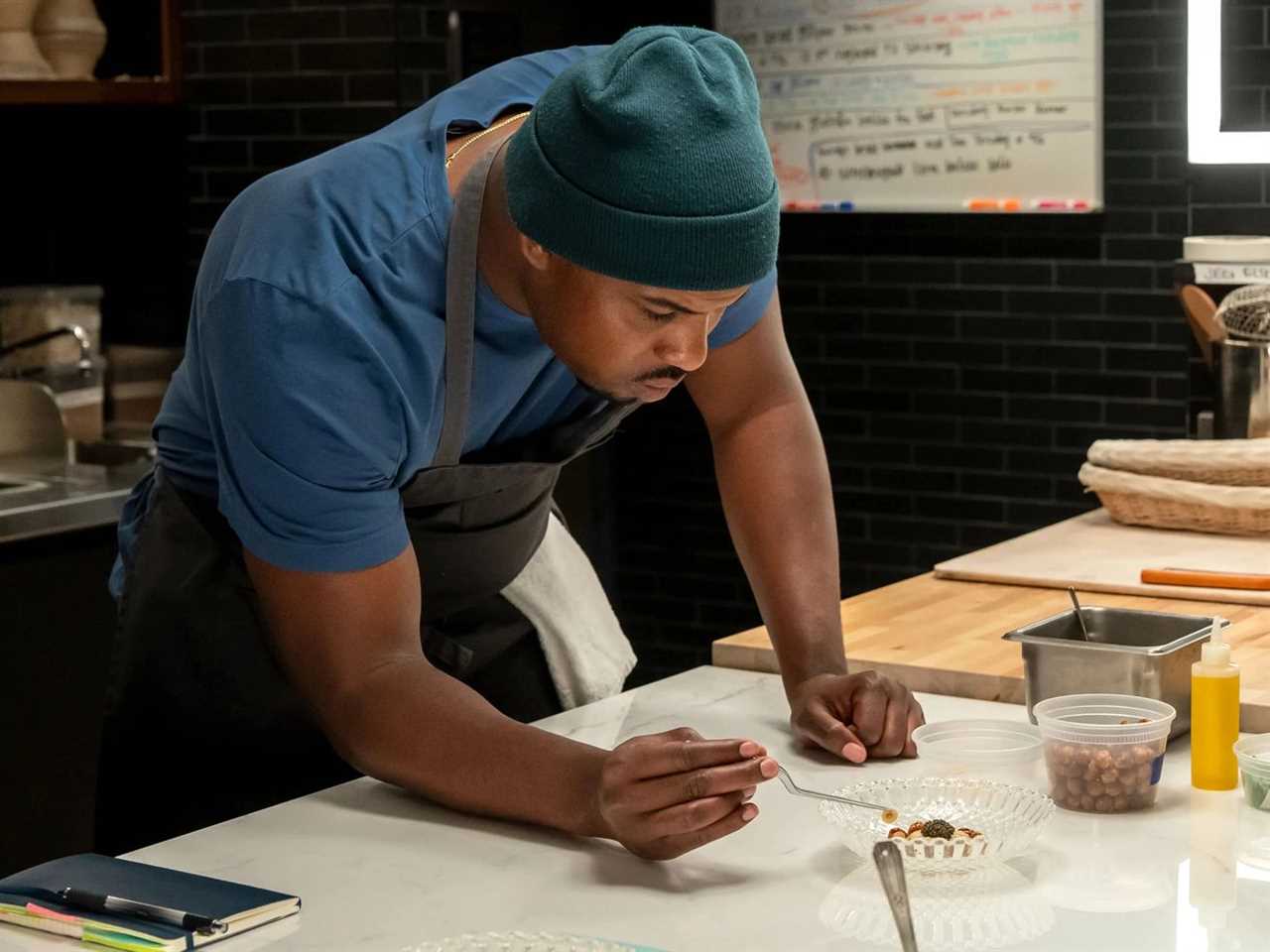 A man in an apron leans over a kitchen prep counter to place a garnish on a dessert.