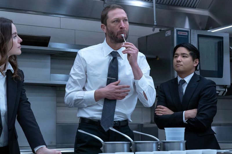 A man in a shirt and tie with a tasting spoon in his mouth is watched by two others in suits flanking him in a restaurant kitchen.