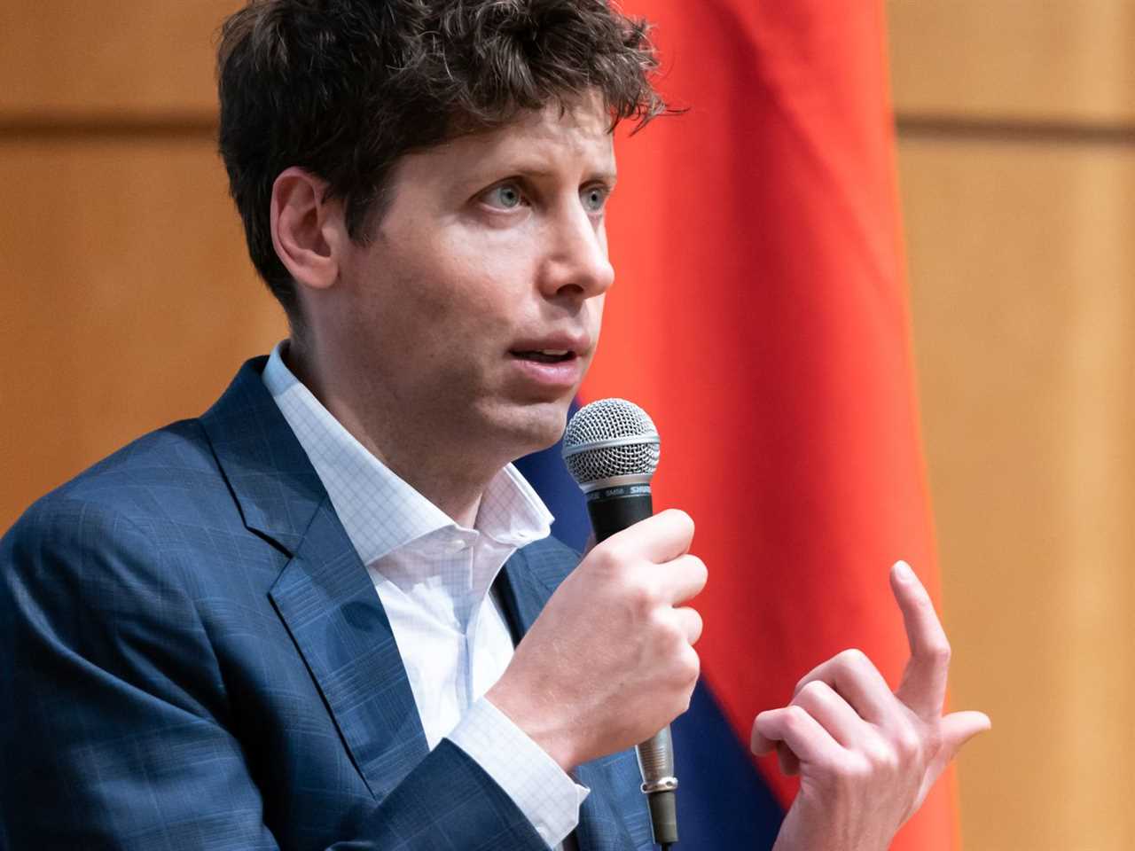 Sam Altman, a white man with curly brown hair wearing a blue suit and white shirt, speaks into a microphone to an unseen audience.